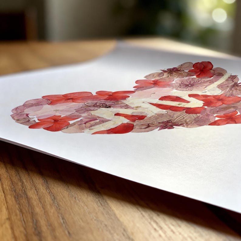 May include: A white rectangular print featuring a heart shape composed of various pressed flowers in shades of red, pink, and muted mauve. The floral arrangement is densely packed, creating a textured heart silhouette on a clean white background.
