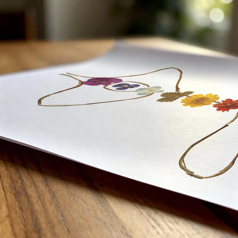 May include: A white art print featuring a line drawing of a human torso silhouette created with pressed flowers. The floral arrangement includes purple, blue, yellow, and red petals along with green leaves, forming the outline of the body shape.