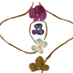 May include: A decorative arrangement of pressed botanical elements featuring a magenta flower, a deep purple flower, and a pale blue flower centered within a frame of thin, dried brown twigs, with a textured green leaf positioned at the bottom.