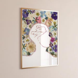 Puede incluir: Una pieza de arte mural rectangular en un marco dorado fino con fondo blanco y el contorno central de un perfil humano. La silueta está rellena y rodeada por un arreglo de flores secas prensadas en tonos morados, azules, blancos y rosados.