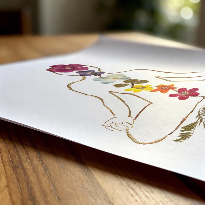 May include: A white art print featuring a line drawing of a human torso outlined in brown. The center of the torso is decorated with a vertical arrangement of pressed, dried flowers in shades of magenta, purple, light blue, green, yellow, and orange.
