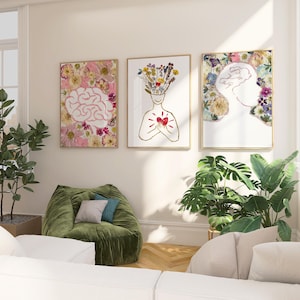May include: Three framed wall art prints featuring botanical illustrations. The left print shows a brain surrounded by pink and yellow flowers. The center print depicts a figure with a floral head and a red heart. The right print shows a floral silhouette.