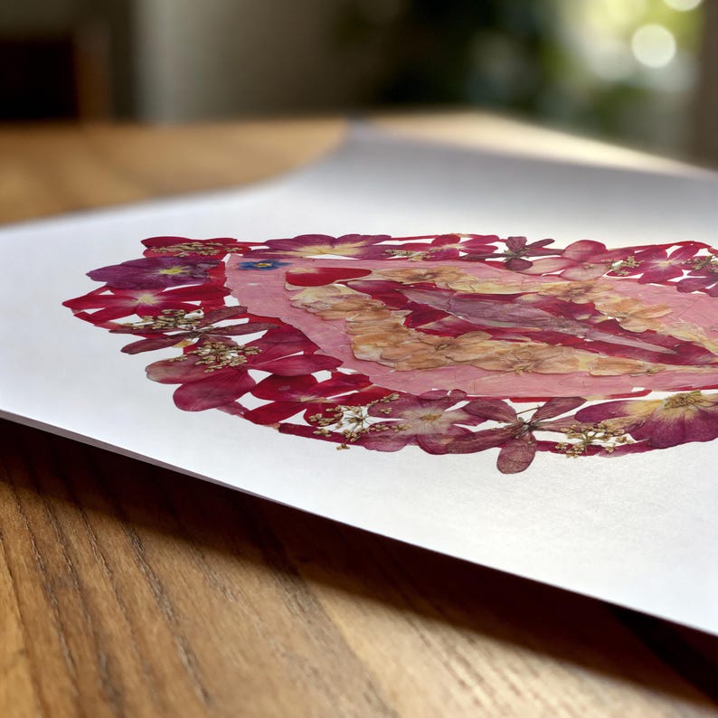K&ouml;nnte beinhalten: Ein wei&szlig;er Kunstdruck mit einer zentralen ovalen Anordnung aus gepressten Blumen in den Farben Magenta, Pink und Creme. Die botanische Komposition ist dicht geschichtet und zeigt verschiedene Bl&uuml;tenblattformen und Texturen.