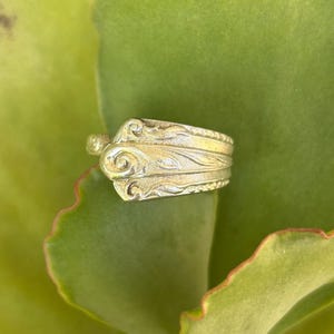 May include: A silver ring made from a repurposed spoon with an ornate design. The ring is on a green succulent plant.