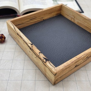 May include: A wooden dice tray with a black felt lining. The tray has a natural wood finish with dark brown streaks. There are several red and gold dice scattered on a white gridded surface.