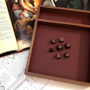 May include: A wooden dice tray with a burgundy felt lining. The tray has a small compartment for storing dice. There are eight metal dice in the tray, including a d20, d12, d10, d8, d6, and three d4 dice.