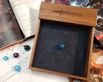 May include: A wooden dice tray with a dark blue interior and a decorative sword and dice design on the lid. A blue and gold twenty-sided die rests inside the tray. Other dice and role-playing game books are visible.