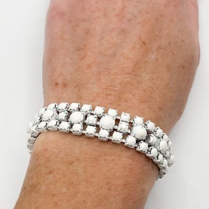 May include: A silver-toned metal bracelet featuring three parallel rows of clear, square-cut rhinestones interspersed with round, opaque white cabochon stones. The bracelet has a flexible, multi-strand design that wraps around the wrist.