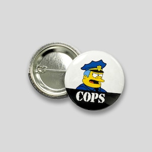 May include: A white button with a black and white image of a yellow cartoon character wearing a blue police hat and the word "COPS" in white letters on a black background.