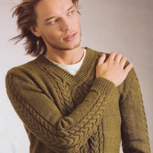 以下が含まれることがあります： Vネックのオリーブグリーンのケーブルニットセーター。セーターは前面と袖に複雑なケーブル模様が施されています。「Mr Buntings Coat」の文字が右上に見えます。白いアンダーシャツがセーターの下に見えます。