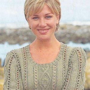 May include: A light green knitted top with short sleeves. The top features a round neckline and a textured pattern with vertical and diamond-shaped details. The sleeves have a decorative openwork design. The woman is smiling.