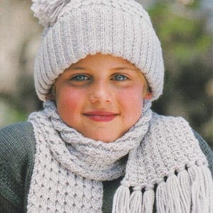 Könnte beinhalten: Ein hellgraues Strickmützen- und Schal-Set mit Fransen. Die Mütze ist im Beanie-Stil gehalten und der Schal hat ein strukturiertes Muster. Die Person trägt einen dunkelgrünen Pullover. Dieses gemütliche Set ist perfekt für kaltes Wetter.