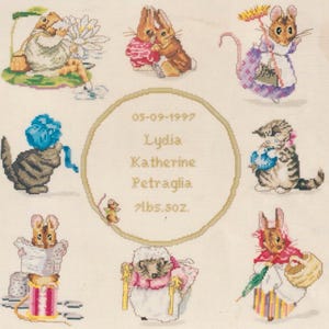 May include: A cross-stitch sampler featuring various animal characters, including rabbits, cats, and mice, in a whimsical design. The sampler includes the text "05-09-1997 Lydia Katherine Petraglia 7lbs.5oz." in a central circle, indicating a birth announcement.