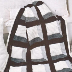 Puede incluir: Una manta de ganchillo hecha a mano con un patrón geométrico. La manta presenta grandes cuadrados en blanco, gris y marrón oscuro, creando un contraste visual llamativo. La manta está drapeada sobre una superficie blanca, mostrando su textura y diseño.