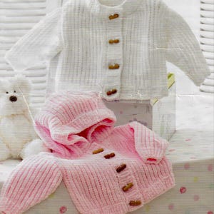 May include: Two baby sweaters. One is white with wooden buttons, the other is pink with a hood and wooden buttons. The sweaters are made of a ribbed knit fabric. The text "Mr Buntings Coat" is visible in the bottom right corner.