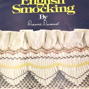 May include: A book cover with the title "The Art Of English Smocking" in yellow letters on a dark blue background. The cover features a close-up of white fabric with detailed smocking in various colors.