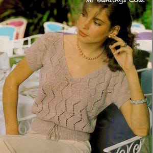 Puede incluir: Una blusa de punto marrón claro con cuello en V y mangas cortas. La blusa presenta un patrón en zigzag y una cintura con cordón. El texto "Mr Buntings Coat" está en la esquina superior derecha. La blusa se combina con pantalones claros.