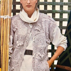 May include: A light beige, speckled knit sleeveless cardigan featuring a button-front closure with four white, elongated toggle-style buttons. The garment has a textured, chunky knit pattern with decorative bobble accents and two front pockets.