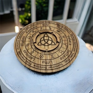 Puede incluir: Un disco de madera redondo con texto y símbolos grabados relacionados con la Rueda del Año. El disco presenta los nombres de las celebraciones estacionales y símbolos astrológicos. El diseño central es un nudo celta.