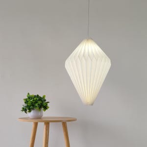 May include: A white, diamond-shaped pendant light with a pleated, paper-like texture hanging from a thin cord. The lamp emits a soft, diffused glow, creating a geometric silhouette that tapers at both the top and the bottom of the fixture.