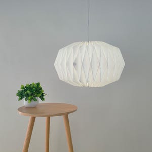 May include: A white, pleated, geometric pendant lamp hangs from a thin cord. Below the lamp, a small, round, light-colored wooden table with three legs holds a small potted green plant with rounded leaves in a white ceramic container.