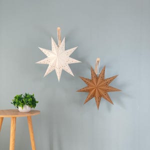 May include: Two paper star decorations, one white and one brown, hang against a light blue wall. The stars have pointed edges and small cut-out patterns. A small potted plant sits on a wooden side table.