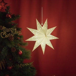May include: A white paper star lantern with a white string hanging from the top. The star has a cut-out design with smaller stars inside. The star is hanging against a red background.