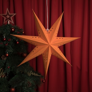 May include: A large, orange paper star with a seven-point design, suspended by a white string. The star has small, star-shaped cutouts. A Christmas tree with red ornaments and a star tree topper is in the background.