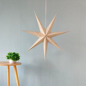 Puede incluir: Una gran estrella de papel beige con ocho puntas cuelga de una cuerda blanca. La estrella tiene pequeñas perforaciones espaciadas uniformemente. La estrella está suspendida frente a una pared azul claro. Una pequeña mesa con una planta está en la esquina inferior izquierda.