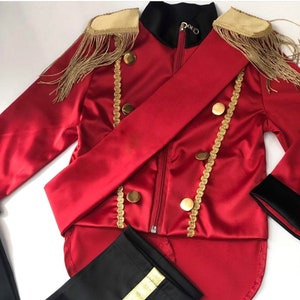 Puede incluir: Una chaqueta de disfraz roja con ribetes dorados, botones y charreteras con flecos dorados. La chaqueta tiene un cuello negro y una faja roja a juego. También se incluye un pantalón negro con una raya dorada.