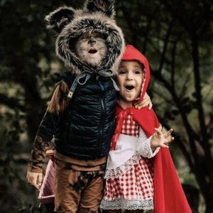 May include: Two children dressed in costumes for Halloween. One child is wearing a wolf costume with a furry hood and brown pants. The other child is wearing a red hooded cape and a red and white checkered dress.