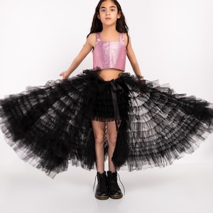 Könnte beinhalten: Ein junges Mädchen trägt ein pinkfarbenes, paillettenbesetztes Crop-Top und einen schwarzen Tüllrock mit hohem/tiefem Saum. Der Rock ist mehrlagig und voluminös, kombiniert mit schwarzen Stiefeln. Der Hintergrund ist weiß.