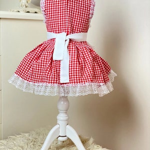 May include: Red and white gingham dress with a white lace trim and a white sash. The dress is on a white dress form.