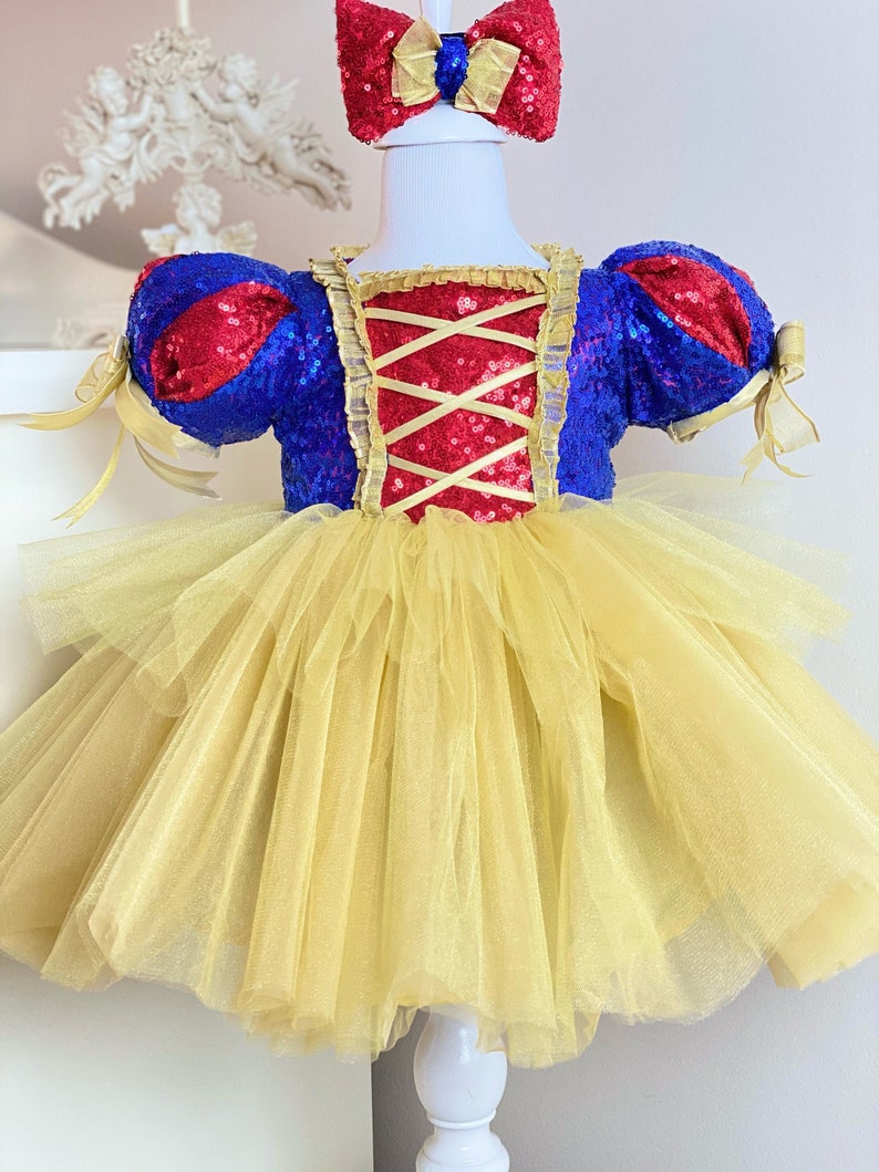 May include: A yellow tulle tutu dress with a red and blue sequined bodice and red bow headband. The bodice has a criss-cross design in the back.