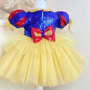 May include: A yellow tulle tutu dress with a blue and red sequined bodice and a red bow. The dress has red and gold ribbon accents and a matching red bow headband.