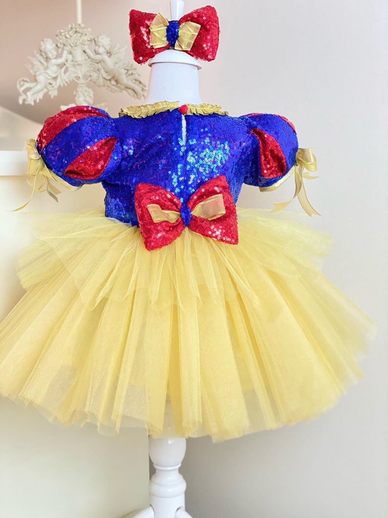 May include: A yellow tulle tutu dress with a blue and red sequined bodice and a red bow. The dress has red and gold ribbon accents and a matching red bow headband.