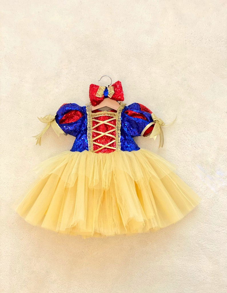 May include: A yellow tulle tutu dress with a red, blue, and gold sequined bodice. The bodice has a criss-cross design and the dress is on a hanger.