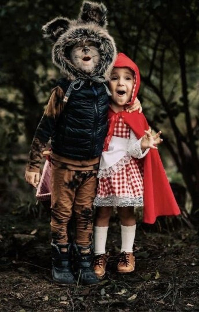 May include: Two children dressed in costumes for Halloween. One child is wearing a wolf costume with a furry hood and brown pants. The other child is wearing a red hooded cape and a red and white checkered dress.