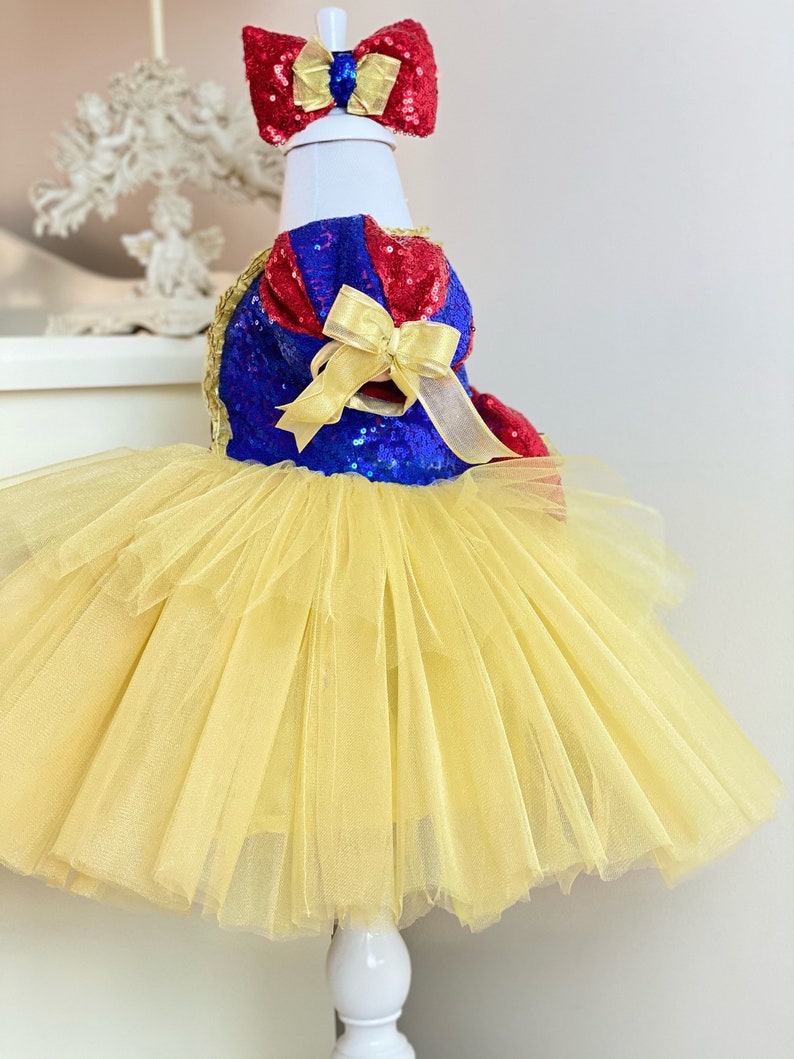 May include: A yellow tulle tutu with a blue and red sequined bodice and a yellow bow. The bodice has a white trim and a red bow on top.