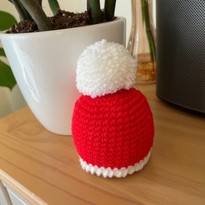 May include: A small, handmade, red and white crocheted Santa hat with a fluffy white pom-pom on top. The hat is sitting on a light-colored wooden surface. The hat is a festive holiday decoration.