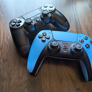 May include: Two game controllers, one black and one blue, are shown on a wooden surface. The blue controller has black accents and coloured buttons. The black controller is positioned above the blue one. Both controllers have multiple buttons and joysticks.