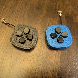 May include: Two square keychains resembling game controller directional pads. One is black, the other blue. Each has a black directional pad and a small silver ring attached to a black cord.