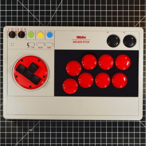 Może przedstawiać: 8BitDo Arcade Stick w jasnoszarym kolorze. Posiada czerwony pad kierunkowy, osiem czerwonych przycisk&oacute;w akcji i dwa czarne przyciski. Na urządzeniu wydrukowano tekst "8BitDo ARCADE STICK".