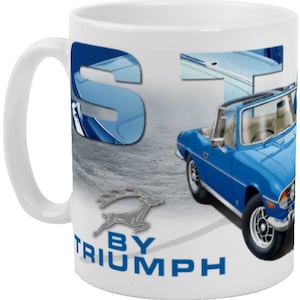 May include: A white ceramic mug with a blue and white image of a Triumph Stag convertible car. The car is blue with a white top and a silver deer logo. The text "BY TRIUMPH" is printed on the mug.