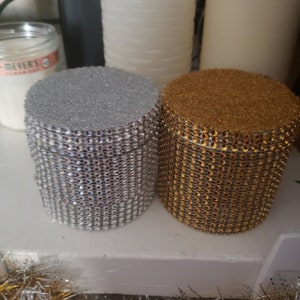 Puede incluir: Dos portavelas decorativos, uno plateado y otro dorado, con un acabado brillante tipo strass. Los portavelas son cilíndricos y tienen una superficie texturizada.