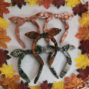 May include: Four fabric headbands with knotted bow details, featuring various patterns including orange plaid, white floral on orange, green plaid, and grey plaid, arranged in a cluster surrounded by colorful autumn maple leaves.