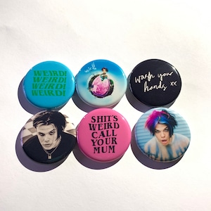 Puede incluir: Seis botones redondos con varios diseños y texto. Los botones presentan frases como "WEIRD!" y "wash your hands", junto con imágenes de personas. La paleta de colores incluye azul, rosa y negro.