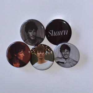 May include: Five round buttons with images of a man with dark hair and a black and white photo of a man with the word "Shawn" printed on it.