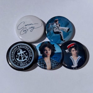 Könnte beinhalten: Eine Sammlung von fünf runden Ansteckbuttons. Ein Button zeigt die handschriftliche Signatur "Conan Gray" auf weißem Hintergrund. Weitere Buttons zeigen Bilder einer Person und den Text "WISHBONE SAILING CLUB".