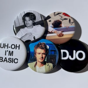 May include: Five circular badges with various designs. One badge displays the text "UH-OH I'M BASIC" in black on white. Other badges feature images of a person, and the letters "DJO" in white on a black background.